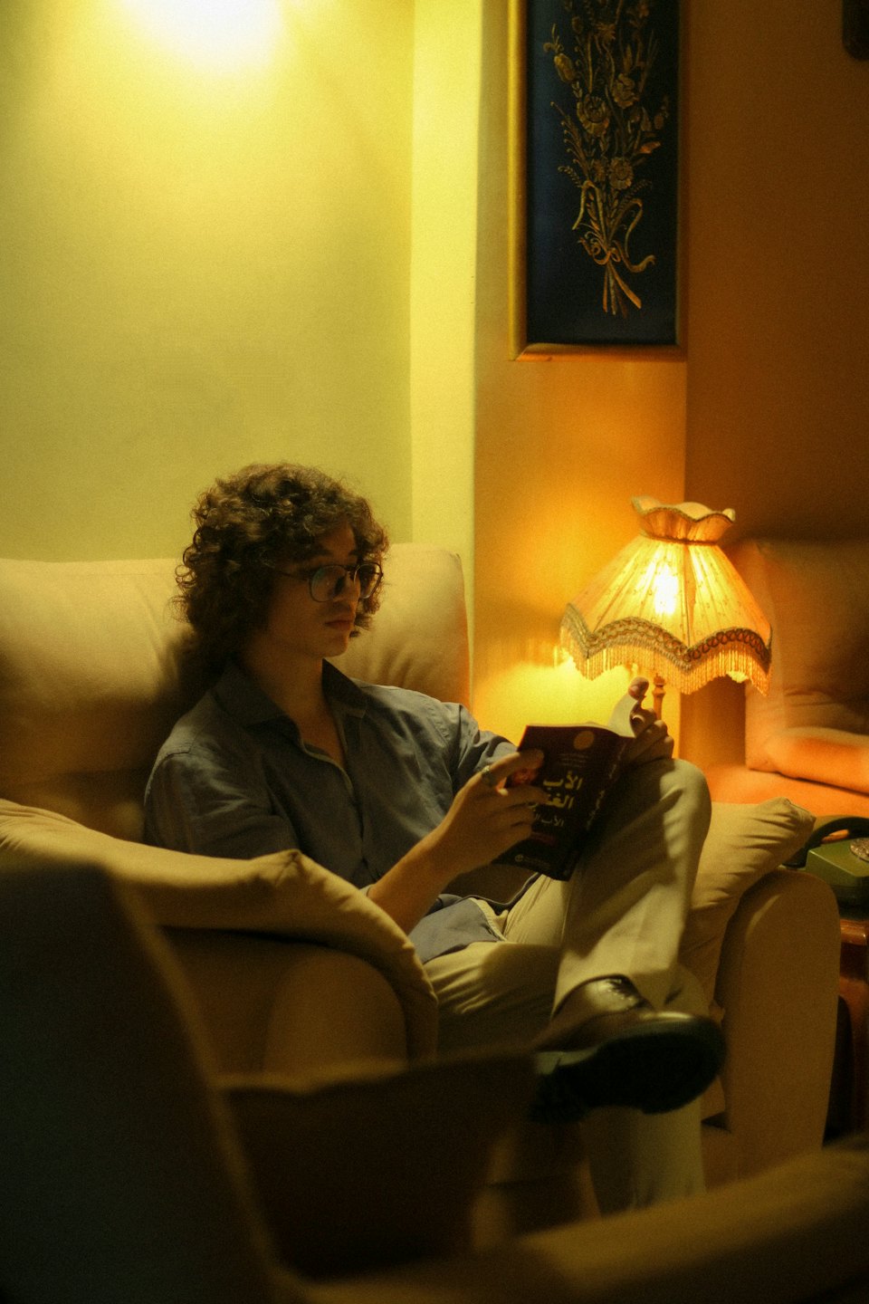 Person with glasses reads a book while sitting in an armchair.